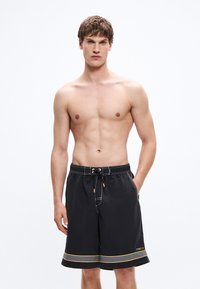 Μαύρο μαγιό βερμούδα με ελαστική μέση και κορδόνι, λευκές ραφές και κίτρινο διακοσμητικό ρίγα στο τελείωμα. Η βερμούδα διαθέτει τσέπες στα πλάγια.