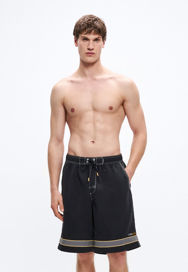 Μαύρο μαγιό βερμούδα με ελαστική μέση και κορδόνι, λευκές ραφές και κίτρινο διακοσμητικό ρίγα στο τελείωμα. Η βερμούδα διαθέτει τσέπες στα πλάγια.