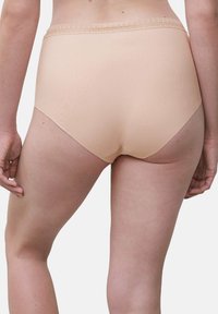 Frau trägt hochtaillierte nahtlose Unterwäsche in Nude mit Spitzenbesatz, von hinten vor einem schlichten hellen Hintergrund gezeigt.