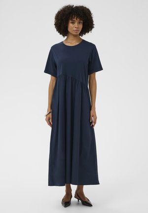 Kobieta o kręconych włosach w granatowej, krótkorękawowej sukience maxi i czarnych, spiczastych sandałach na obcasie, stojąca na tle białego tła.