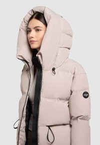 Helepõhjas puffer-jakk, millel on suur kapuuts, must tõmblukk, elastsed köied ja logopatch vasakul varrukal.