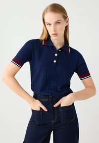 Polo shirt a righe blu navy con pattina a bottoni, bottoni dorati e bordi rossi e bianchi a contrasto sul colletto e sulle maniche. Abbinato a jeans scuri.