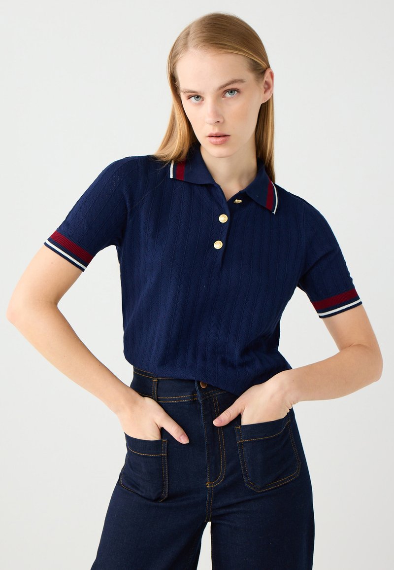 Polo shirt a righe blu navy con pattina a bottoni, bottoni dorati e bordi rossi e bianchi a contrasto sul colletto e sulle maniche. Abbinato a jeans scuri.