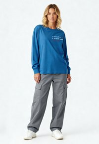 Femme aux cheveux blonds portant une chemise bleue à manches longues avec un texte, un pantalon cargo gris et des baskets blanches, debout devant un fond uni.