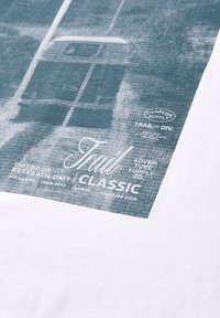 Gros plan sur un tissu avec un graphisme bleu délavé d’un camping-car et un texte blanc incluant « Trail Classic » ainsi que des logos d’équipements de plein air.