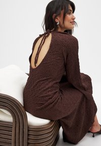 Robe brune texturée avec des manches longues et un dos ouvert, maintenue par un lien. Portée en étant assise sur un fauteuil en courbe brun.