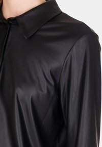 Camicia nera aderente in materiale lucido e liscio con colletto strutturato e pattina con bottoni, caratterizzata da una sottile trama e lucentezza.