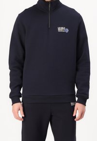 Sudadera con cremallera en color navy, fabricada en un material suave, que cuenta con un cuello alto, puños acanalados y un parche con el logo en el pecho. Combinada con pantalones a juego.