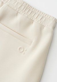 Light beige joggers with an elastic waistband, featuring a back pocket and embossed logo. The fabric appears smooth and soft.