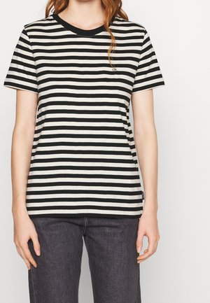 Žena nosící černobílé horizontálně pruhované tričko s krátkým rukávem a malým logem "ck" na prsou, v kombinaci s tmavě šedými džínami.