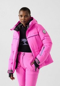 Donna che indossa una giacca imbottita rosa acceso e pantaloni abbinati, con una maglia nera con cerniera, in piedi davanti a uno sfondo chiaro e uniforme.