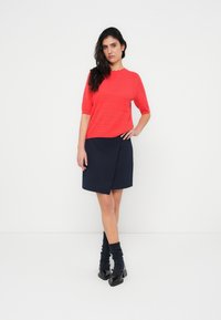 Κόκκινο πλεκτό τοπ με κοντά μανίκια, υφή με ανάγλυφο σχέδιο, σε συνδυασμό με φούστα navy με σχέδιο περιτυλίγματος. Μαύρα παπούτσια με χοντρά τακούνια και σκούροι κάλτσες-ζεστασιά ποδιών ολοκληρώνουν την εμφάνιση.