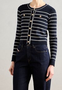 Marineblauwe cardigan met witte strepen, knoopsluiting aan de voorkant en decoratieve knopen; gecombineerd met donkere denim jeans. Zachte, geribbelde textuur.