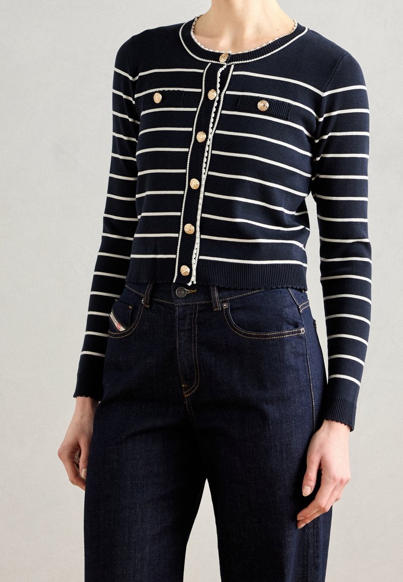 Marineblauwe cardigan met witte strepen, knoopsluiting aan de voorkant en decoratieve knopen; gecombineerd met donkere denim jeans. Zachte, geribbelde textuur.