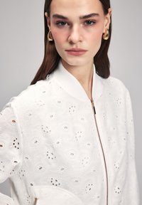 Giacca bomber bianca con design ad occhiello, colletto a coste e zip frontale. Tessuto texturizzato con intricate fantasie ritagliate.