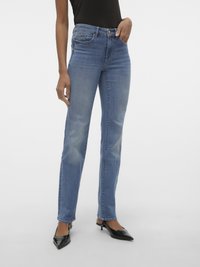 Femme portant un jean droit bleu taille mi-haute avec des décolorations sur les cuisses et des escarpins noirs à bout pointu sur fond blanc.