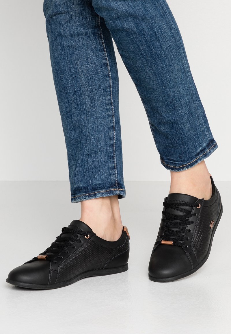 Personne portant un jean bleu foncé et des baskets basses noires décontractées avec des côtés texturés et des lacets noirs sur un sol blanc.