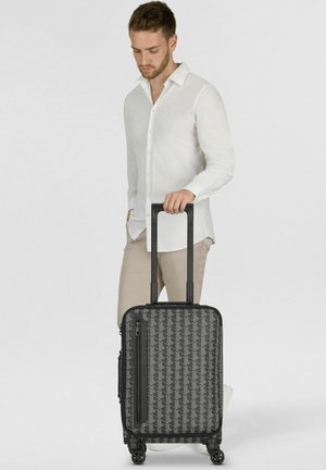 Hombre con camisa blanca y pantalones beige sosteniendo el asa de una maleta negra con estampado y ruedas sobre fondo blanco.