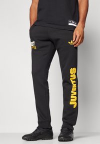 Čierne jogger nohavice vyrobené z jemného materiálu majú na ľavom stehne výrazný žltý text "JUVENTUS" a "1897" a na páse decentné logo.