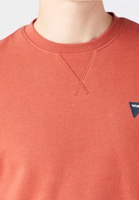 Roestkleurige oranje trui met een ronde halslijn, een gestructureerde ribbelstructuur en een klein marineblauw label met witte tekst in de onderste hoek.