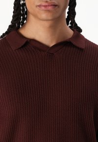 Polo en tricot bourgogne avec une surface texturée, présentant un col et une coupe ample. Les détails incluent des coutures de col inégales.