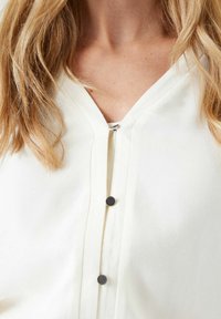 White v-neck top featuring a textured fabric, five small dark buttons down the front, and a relaxed fit with subtle stitching details.