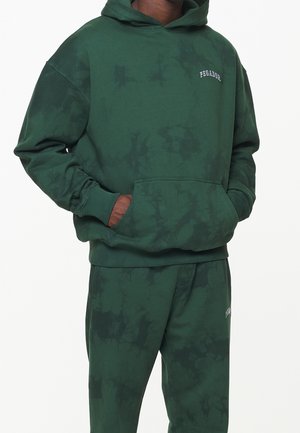 Mann trägt dunkelgrünen Batik-Hoodie und passende Jogginghose mit dem Schriftzug "PEGADOR" auf Brust und Oberschenkel, Hände in der Kapuzen-Tasche.