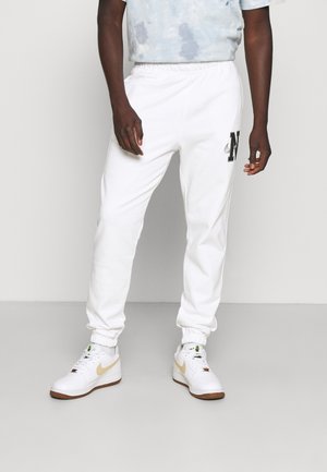 Mann in weißen Nike Jogginghosen mit elastischen Bündchen und einem schwarzen "N"-Logo, kombiniert mit weißen Turnschuhen mit beigen Swooshes.