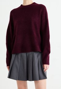 Pull bordeaux en maille avec un ourlet côtelé et des manches longues, associé à une jupe grise évasée et plissée. Texture douce évidente dans le tissu.