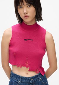 Fuksijas adīta topiņš ar augstu kaklu, ar nolietotām malām un melnu logo. Apvienots ar zilu džinsu šortiem.