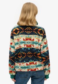 Fleece pullover with a colorful geometric pattern in shades of blue, red, and cream. Features a high collar and green cuffs.