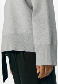 Grå strikket sweater med ribbede manchetter og kant, der har et splitdesign og blød tekstur, båret over sorte bukser.