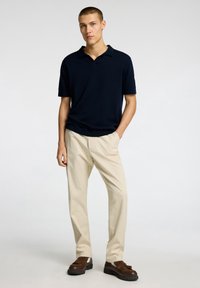 Navy short-sleeve polo shirt paired with beige trousers and brown lace-up shoes. The shirt features a classic collar and relaxed fit.