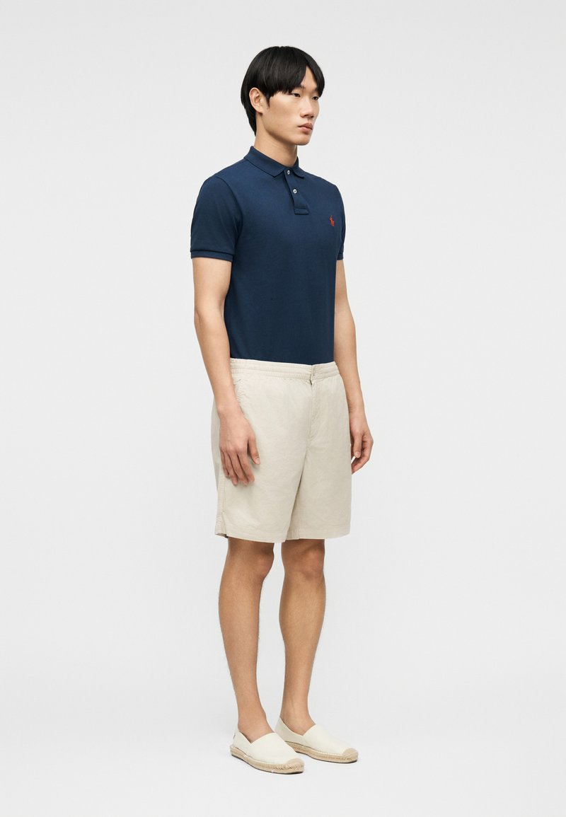 Homme debout, légèrement tourné vers la droite, portant un polo bleu marine, un short beige et des chaussures crème sans lacets, sur un fond uni.
