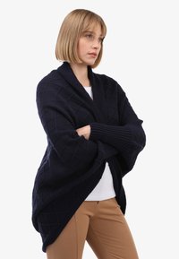 Marineblauer gestrickter Schal-Kardigan mit einem Rautenmuster, weiten Ärmeln, offener Vorderseite und gerippten Bündchen, kombiniert mit einem weißen Oberteil und beigen Hosen.