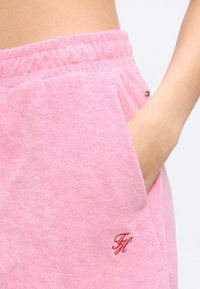 Prim-plan cu mâna unei persoane în buzunarul pantalonilor scurți roz din material tip prosop, cu talie elastică și logo roșu brodat.