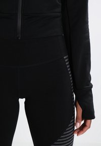 Czarna sportowa kurtka z zippem z przodu, w połączeniu z czarnymi legginsami z teksturowanym paskiem po bokach. Miękka w dotyku tkanina.