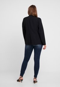 Mujer de pie de espaldas, vistiendo una chaqueta negra, jeans ajustados oscuros y tacones negros de tipo slingback, sobre un fondo gris liso.