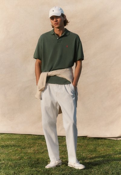 Giovane uomo che indossa una polo verde, pantaloni bianchi, maglione crema legato in vita, cappello bianco e scarpe da ginnastica bianche in piedi sull’erba.