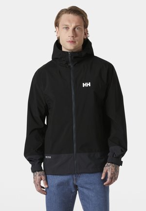Mężczyzna z tatuażami nosi czarną kurtkę z kapturem marki Helly Hansen i niebieskie dżinsy, stoi na tle jednolitego jasnoszarego tła.