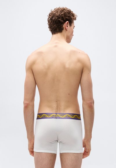 Άνδρας μοντέλο εμφανίζεται από πίσω φορώντας λευκά μπόξερ με διακοσμητική μωβ και χρυσή μέση, σε απλό φόντο.