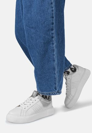 Białe casualowe trampki z perforacjami po bokach, noszone z niebieskimi dżinsami i czarno-białymi skarpetkami w wzory.