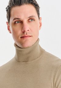 Młody mężczyzna z krótkimi ciemnymi włosami, ubrany w beżowy sweter z golfem, lekko spogląda na bok na tle jednolitego białego tła.