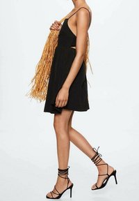 Czarna sukienka bez rękawów z rozkloszowanym dołem, z wycięciami po bokach, połączona z czarnymi sandałami na obcasie. Materiał wydaje się lekki; sukienka ma gładką fakturę.