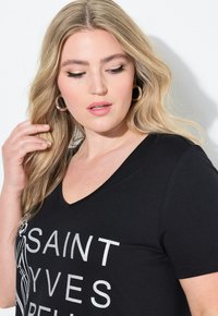 Černé tričko s výstřihem do V a krátkými rukávy, se bílým grafickým textem "SAINT YVES", vyrobené z měkkého materiálu.