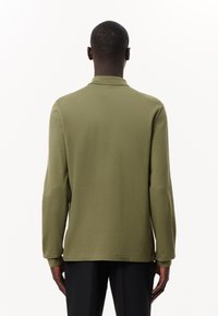 Camicia a maniche lunghe verde oliva con colletto polo, texture liscia e taglio dritto. Presenta spacchi laterali e un orlo pulito.