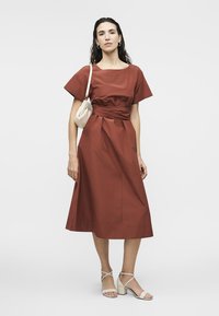 Γυναίκα που στέκεται φορώντας ένα μεσαίο φόρεμα σε σκουριά χρώματος με φαρδιά μανίκια, μπεζ πέδιλα με τακούνι και κρατώντας μια μικρή μπεζ τσάντα ώμου.