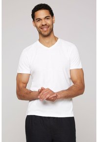 Camiseta blanca de cuello en V hecha de tela suave. Mangas cortas, diseño ajustado. El modelo tiene un complexión media con cabello corto y rizado. El modelo posa con las manos juntas.