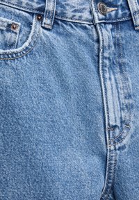 Gros plan d’un jean en denim bleu clair montrant un bouton en métal, des passants de ceinture, une poche avant et des coutures détaillées.