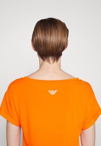 Person med kort brunt hår som bär en ljusorange skjorta med en liten vit prickig logotyp på övre ryggen mot en enkel bakgrund.